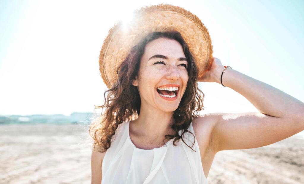 A woman with curly hair smiles broadly while holding a straw hat on her head, standing outdoors on a sunny beach.