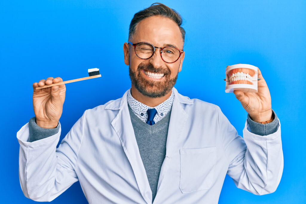 A male dentist with a beard and glasses wearing a white coat, winking while holding a toothbrush and a model of a jaw on a blue background.