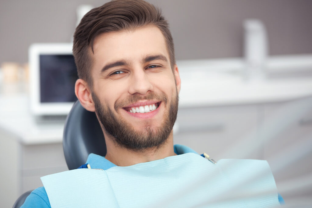 Portrait of a smiling young man with a beard sitting in a dental chair. He is wearing a blue bib and looking directly at the camera with a happy expression